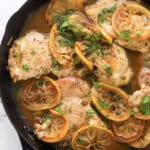 Juicy lemon chicken piccata breasts cooked in a cast-iron skillet with sliced lemons and fresh parsley, coated in a light brown sauce. The dish is garnished with herbs, making it flavorful and visually appealing.
