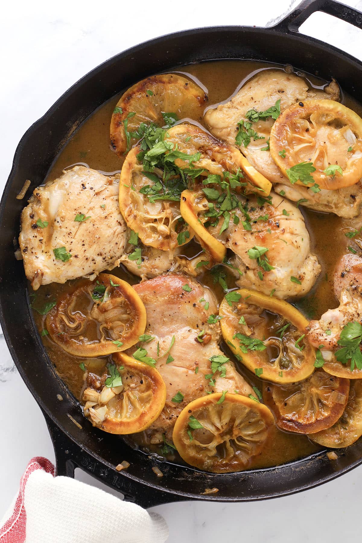 Juicy lemon chicken piccata breasts cooked in a cast-iron skillet with sliced lemons and fresh parsley, coated in a light brown sauce. The dish is garnished with herbs, making it flavorful and visually appealing.