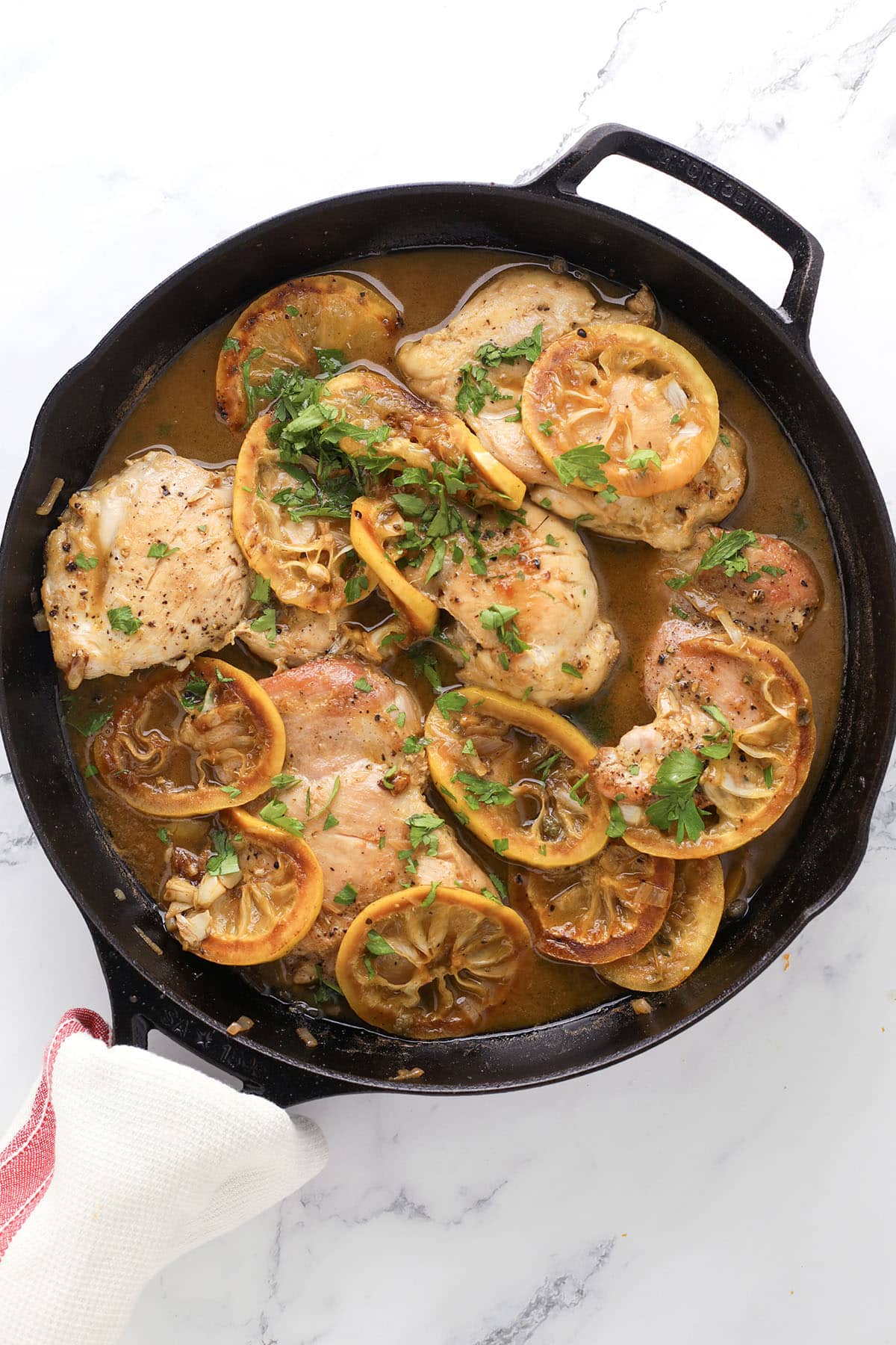 A cast iron skillet with lemon chicken piccata—tender chicken breasts topped with lemon slices and fresh chopped herbs in a brown sauce—set on a white marble surface.