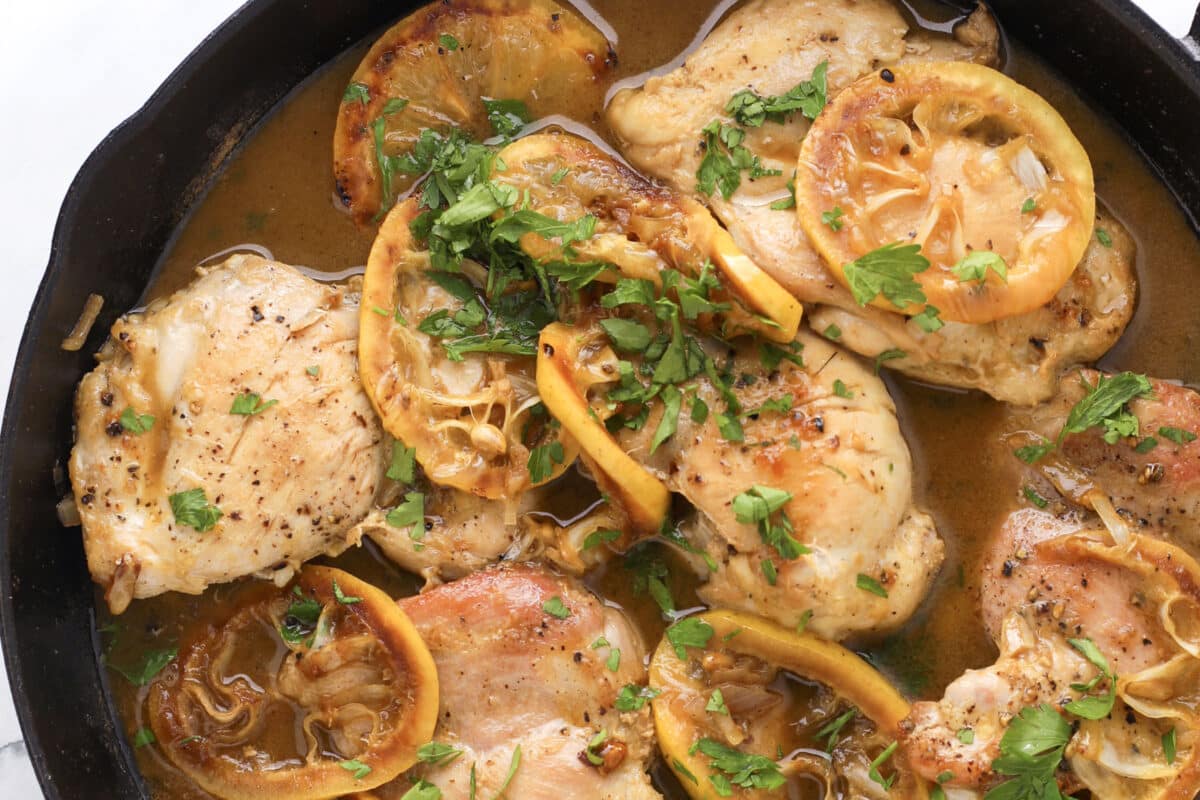 Chicken thighs cooked in a skillet with lemon slices, fresh parsley, and a savory sauce—this take on lemon chicken piccata is garnished with chopped herbs and surrounded by a rich, flavorful broth.