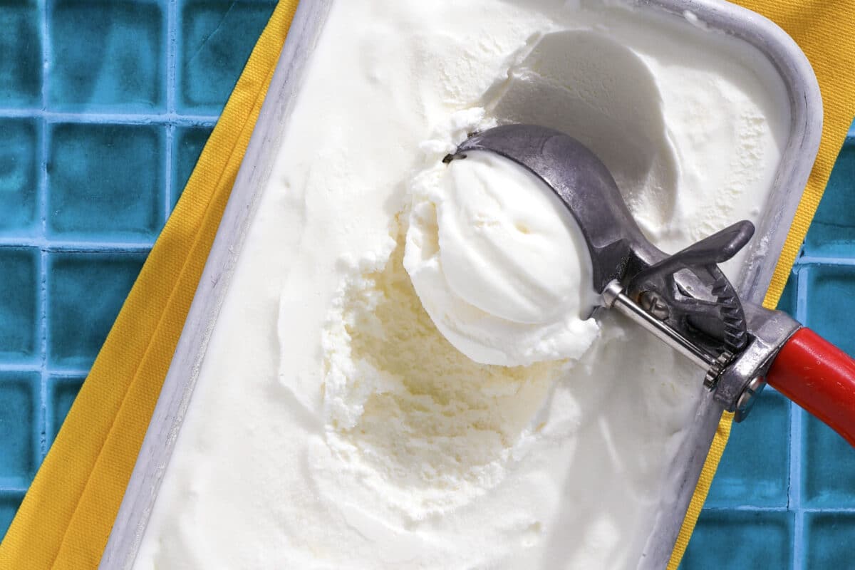 A metal ice cream scoop with a red handle is serving homemade lemon ice cream from a rectangular container, which sits on a yellow cloth and blue tiled surface.
