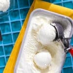 A tub of lemon ice cream with two scoops and a metal ice cream scooper rests on a yellow napkin atop a blue tiled surface. A bowl with a scoop of ice cream is partially visible nearby.
