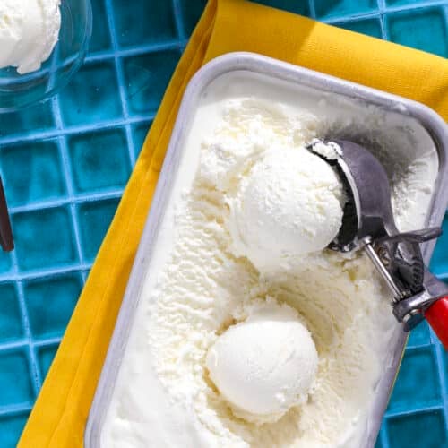 A tub of lemon ice cream with two scoops and a metal ice cream scooper rests on a yellow napkin atop a blue tiled surface. A bowl with a scoop of ice cream is partially visible nearby.