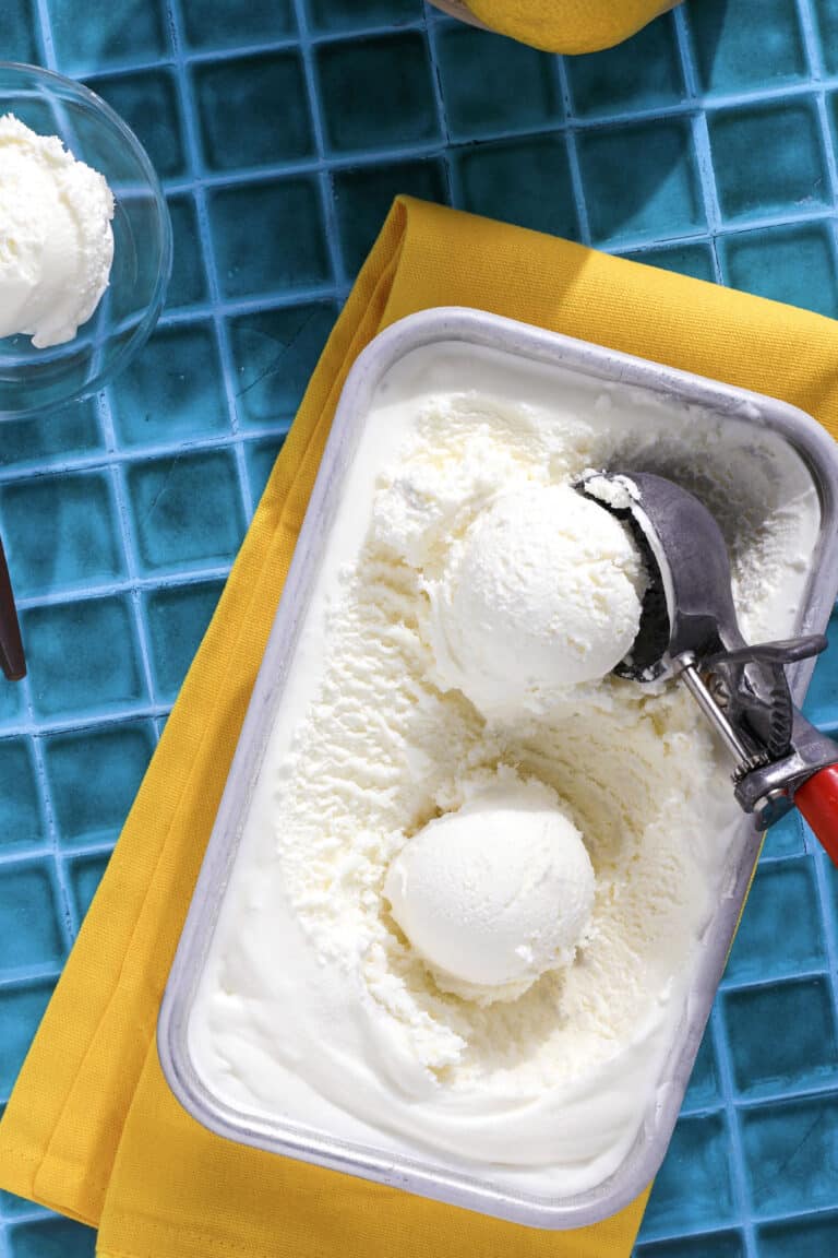 A tub of lemon ice cream with two scoops and a metal ice cream scooper rests on a yellow napkin atop a blue tiled surface. A bowl with a scoop of ice cream is partially visible nearby.