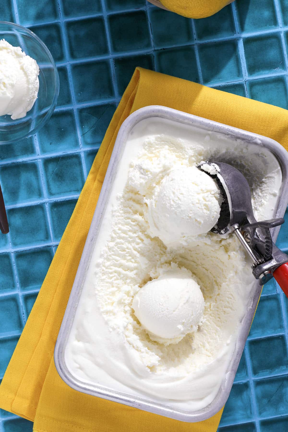 A tub of lemon ice cream with two scoops and a metal ice cream scooper rests on a yellow napkin atop a blue tiled surface. A bowl with a scoop of ice cream is partially visible nearby.