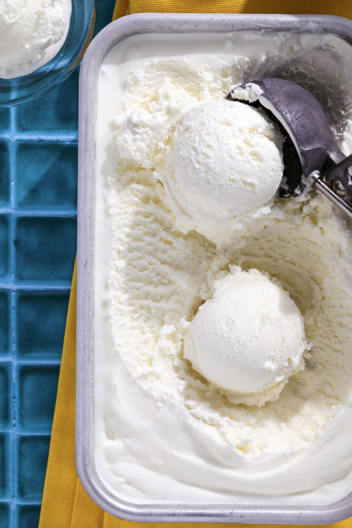 A metal ice cream scoop serves vanilla and easy lemon ice cream from a rectangular container, with two scoops in the container and a bowl of ice cream nearby on a blue tiled surface with a yellow cloth.