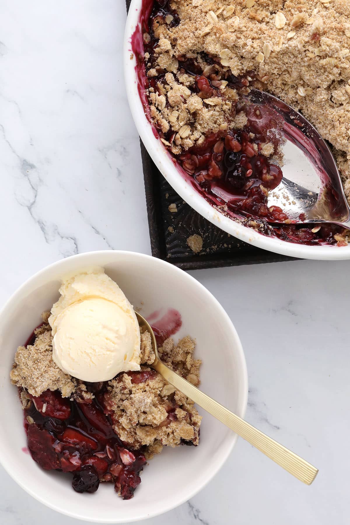 A bowl with mixed berry crisp and a scoop of vanilla ice cream sits beside a baking dish filled with more berry crisp, complete with a serving spoon. The dessert rests on a white marble surface.