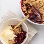 A bowl and a baking dish with mixed berry crisp topped with vanilla ice cream sit on a beige napkin. A spoon is in the bowl, and part of the crisp has been served from the baking dish.
