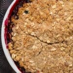 A close-up of a baked fruit crumble in a white dish, topped with a golden oat and crumb topping. The fruit filling beneath is dark and juicy, with some of it bubbling up around the edges.