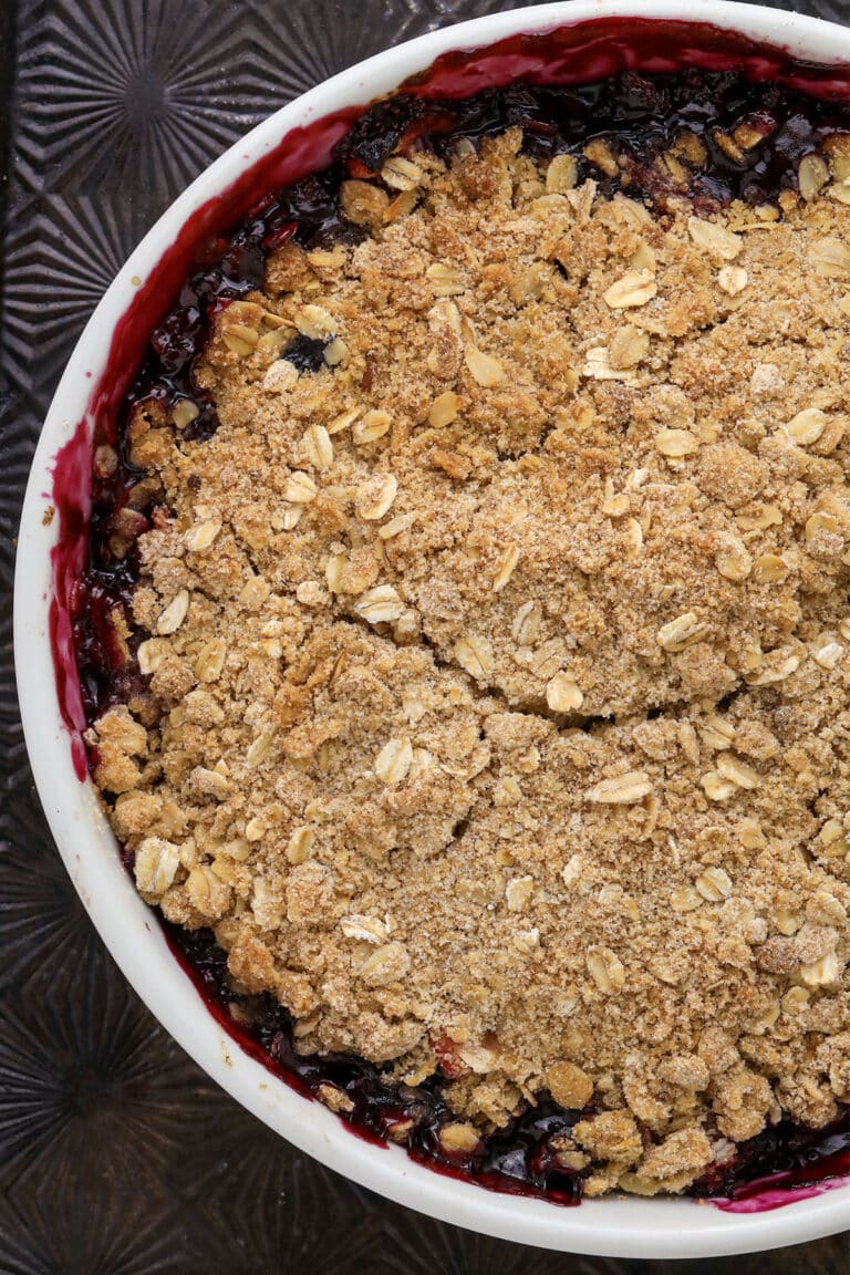 A close-up of a baked fruit crumble in a white dish, topped with a golden oat and crumb topping. The fruit filling beneath is dark and juicy, with some of it bubbling up around the edges.