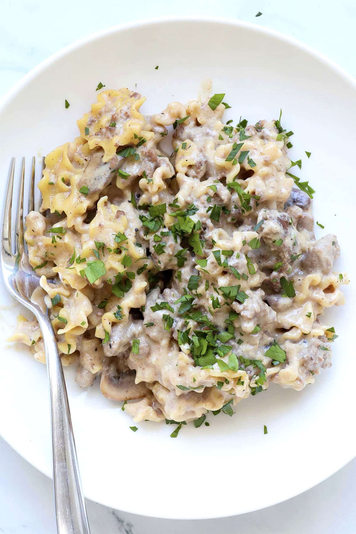 Pasta with creamy mushroom and ground meat sauce, topped with chopped parsley, served on a white plate with a fork.