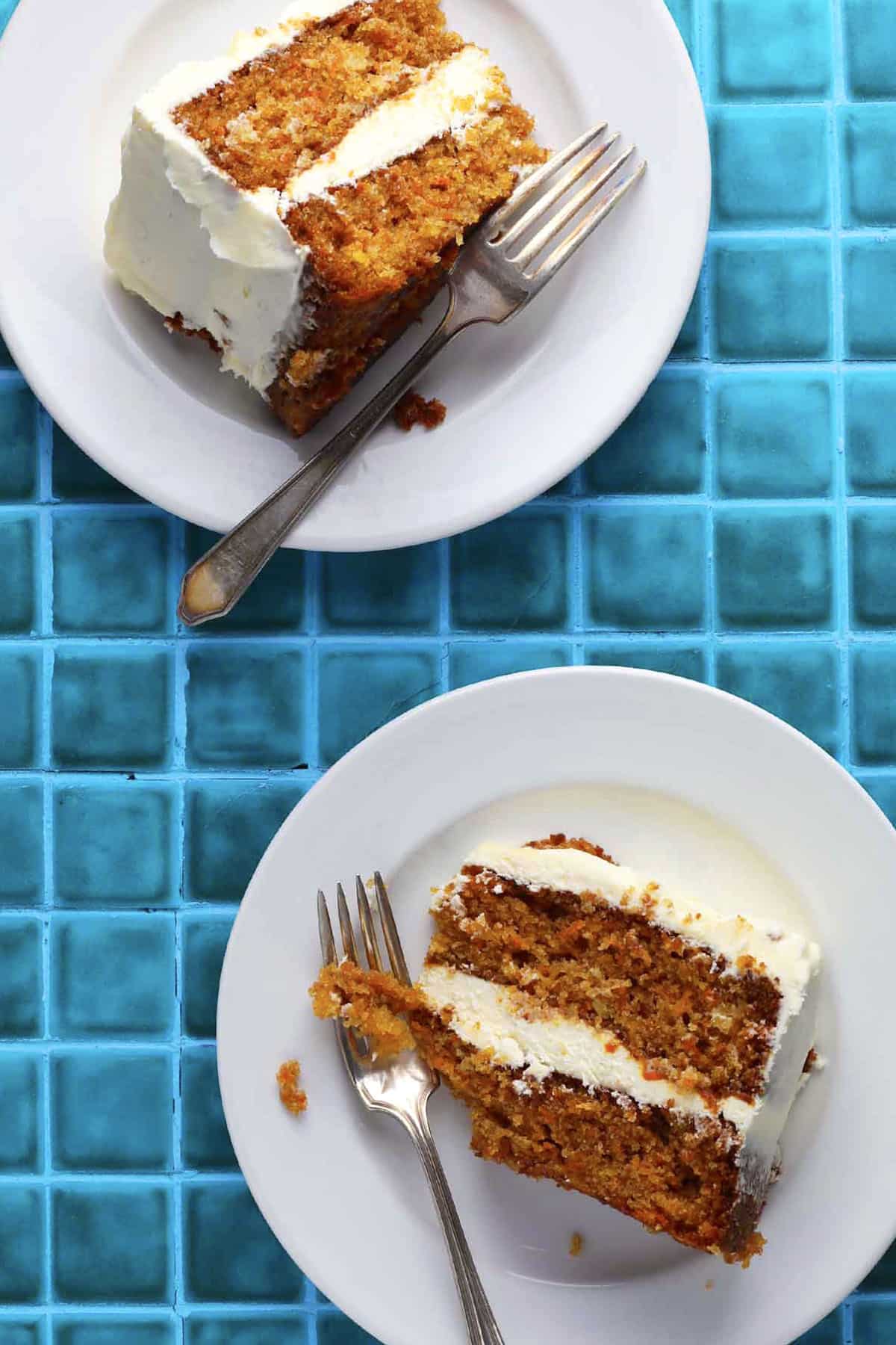 Two plates on a bright blue tiled surface, each with a slice of carrot cake topped with cream cheese frosting. A fork rests on each plate, one piece of cake partially eaten.