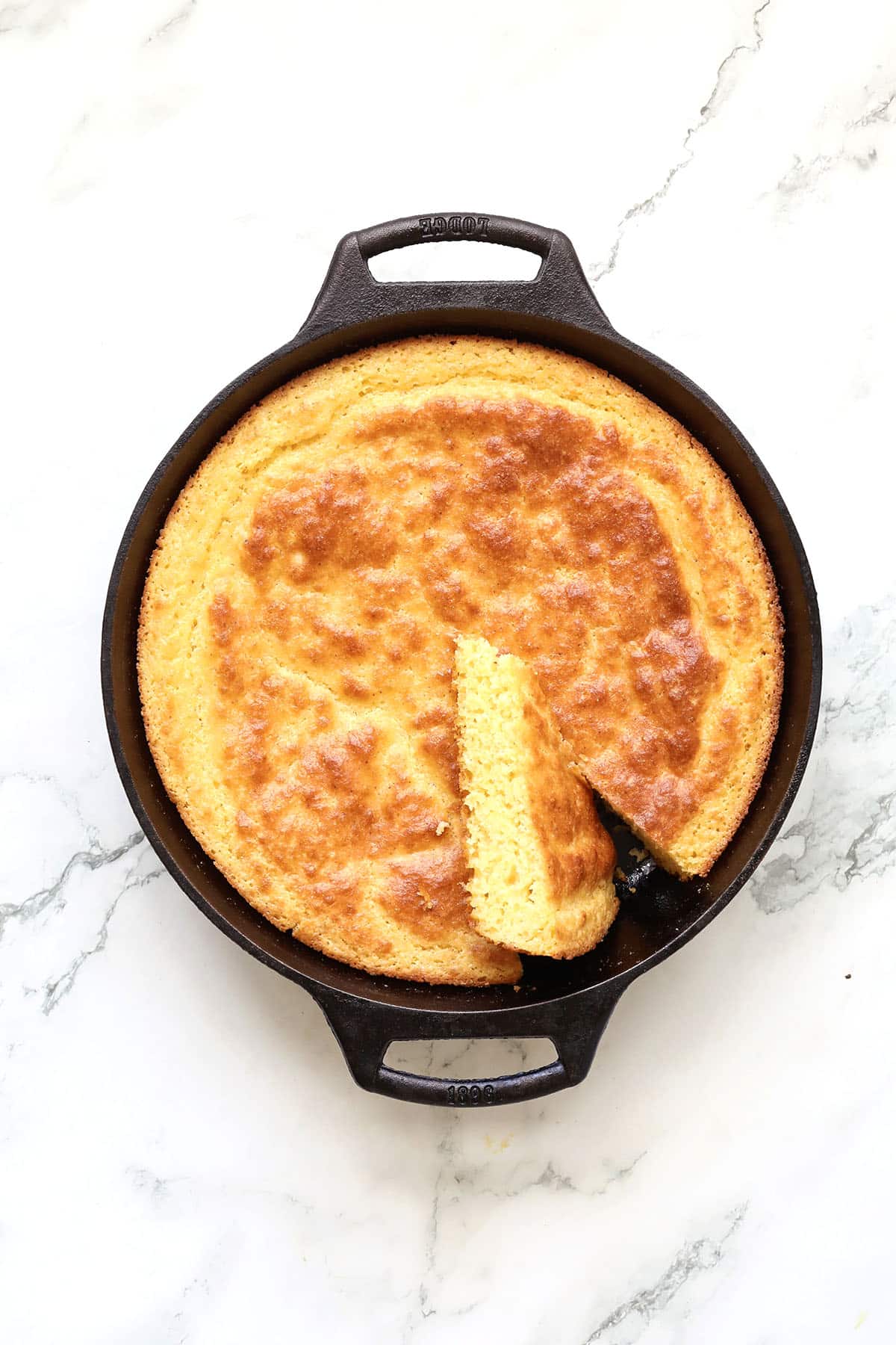 A golden-brown cornbread in a black cast iron skillet with one wedge cut out, viewed from above on a white marble surface.