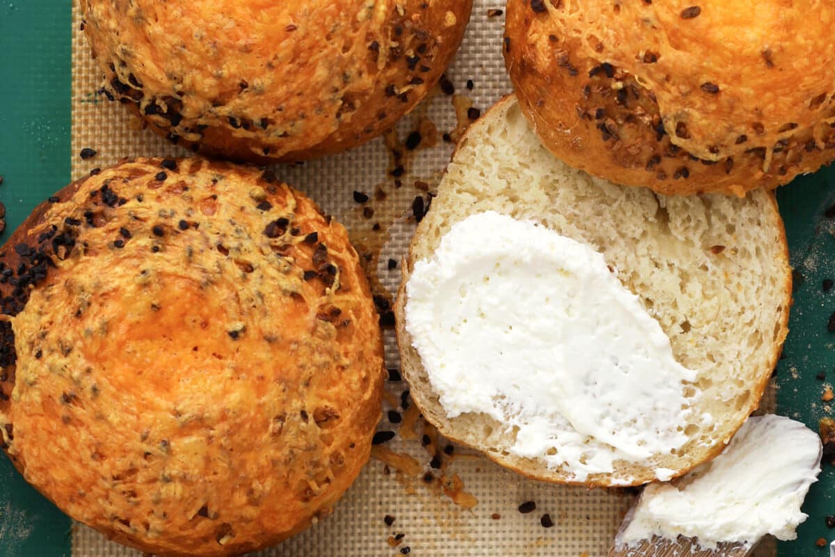 Four golden cheddar garlic bagels on a silicone mat; one is cut open with a thick layer of creamy white cheese spread inside. Crumbs and bits of cheese are scattered around, capturing the delicious results of learning how to make bagels at home.