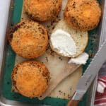 Round, golden brown cheesy garlic bagels sit on a baking tray. One is sliced open and spread with cream cheese using a wooden spatula. A bread knife and red-striped towel are nearby, adding to the inviting "how to make bagels" scene.