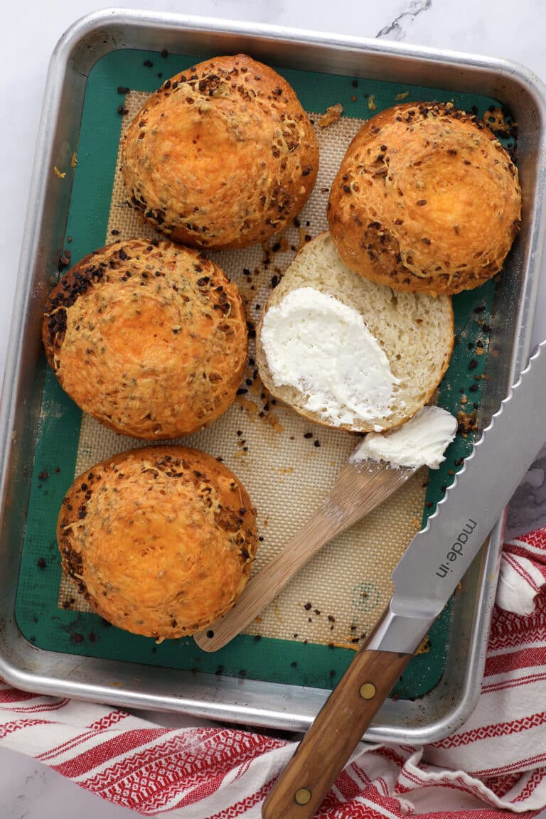Round, golden brown cheesy garlic bagels sit on a baking tray. One is sliced open and spread with cream cheese using a wooden spatula. A bread knife and red-striped towel are nearby, adding to the inviting "how to make bagels" scene.