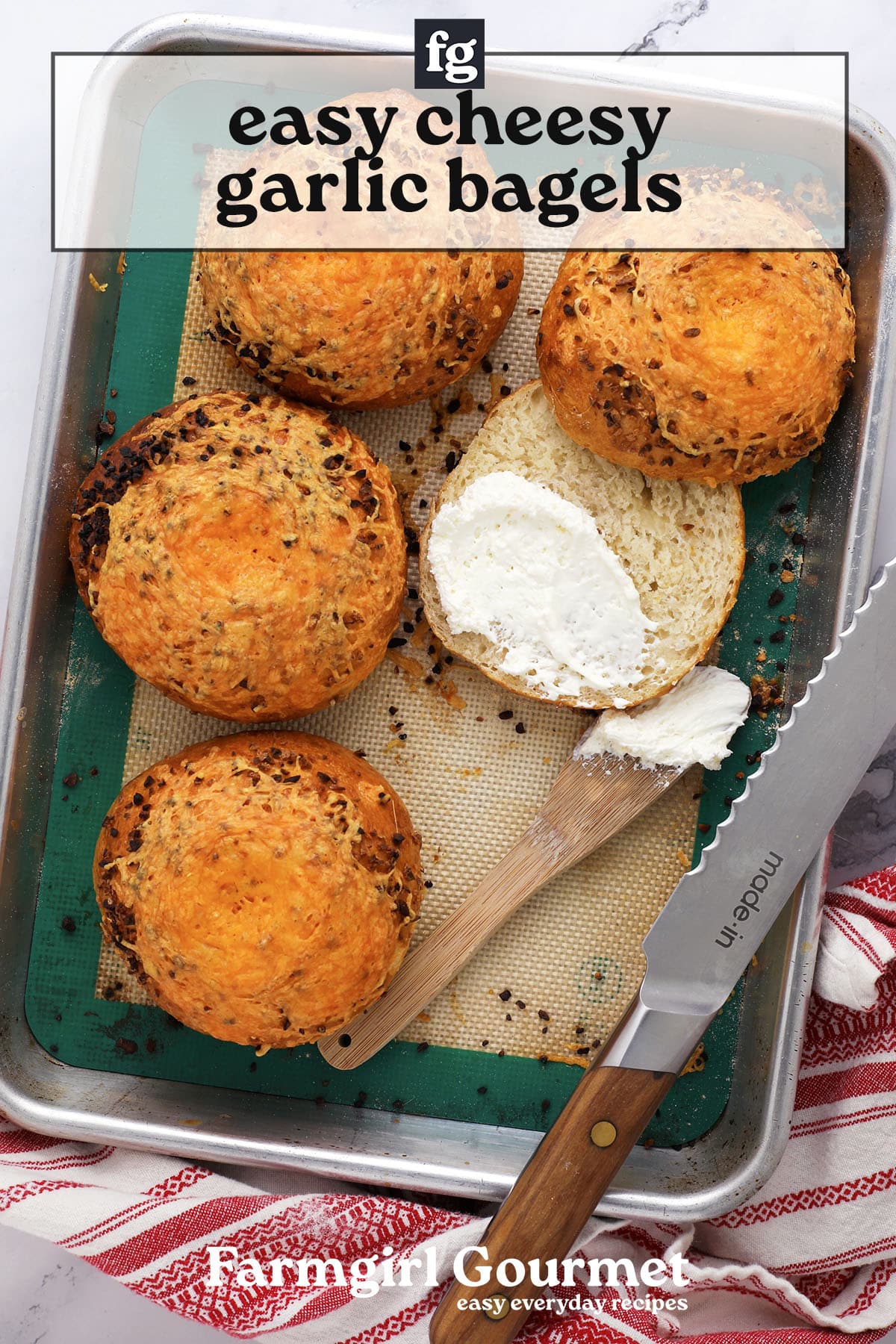 A baking tray with four golden, cheesy garlic bagels. One is sliced open and spread with cream cheese using a wooden knife. A red-striped towel and bread knife are nearby. Text reads easy cheesy garlic bagels—learn how to make bagels at home!.