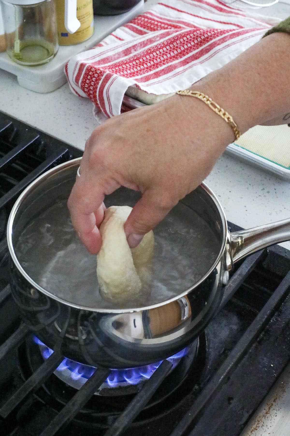 A hand lowers a piece of dough into a pot of boiling water on a stovetop, demonstrating how to make bagels, with a red and white towel in the background.