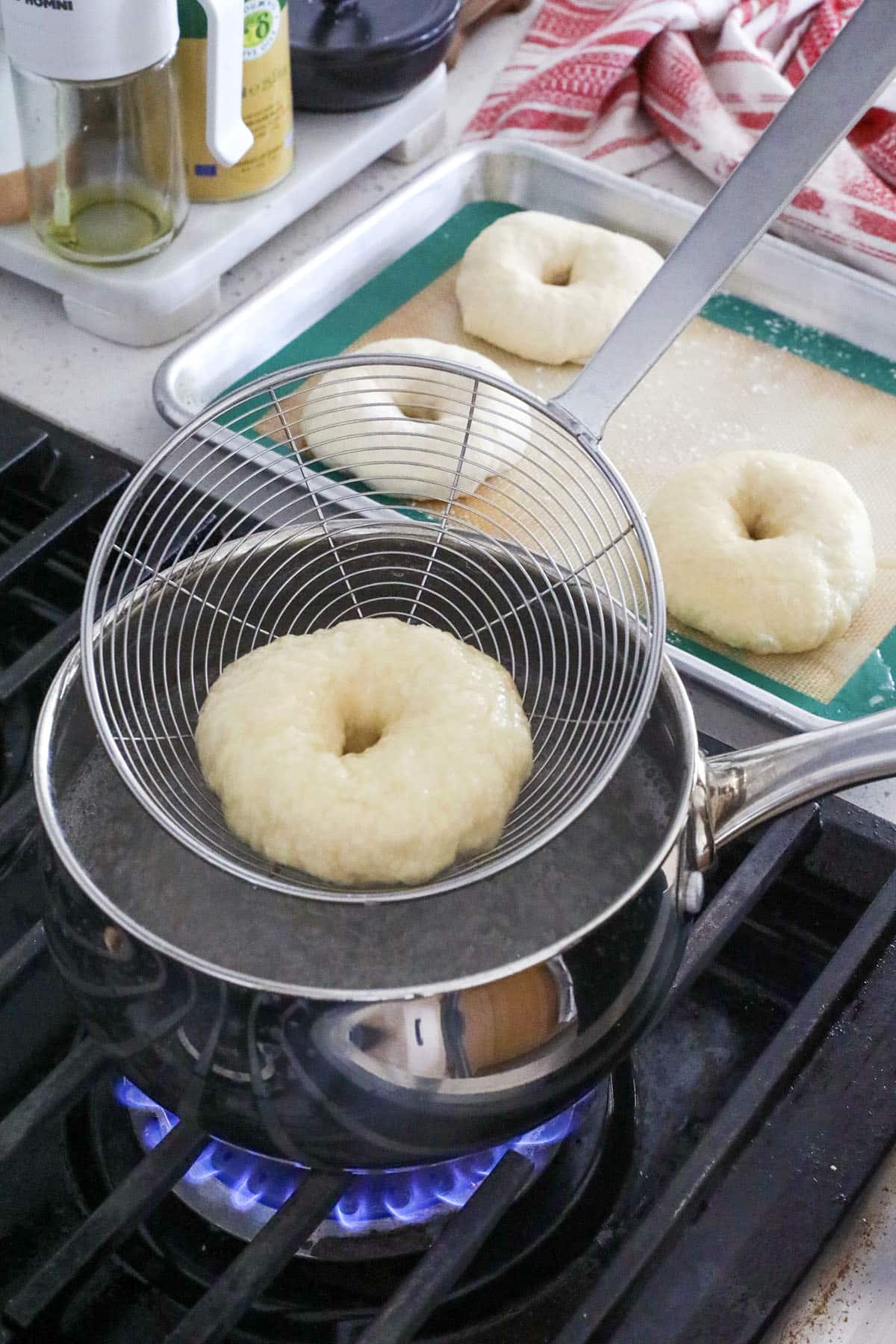 A raw cheddar garlic bagel is being lowered into a pot of boiling water on a stove using a wire skimmer, with more unboiled bagels resting on a baking sheet nearby—showing how to make bagels at home.