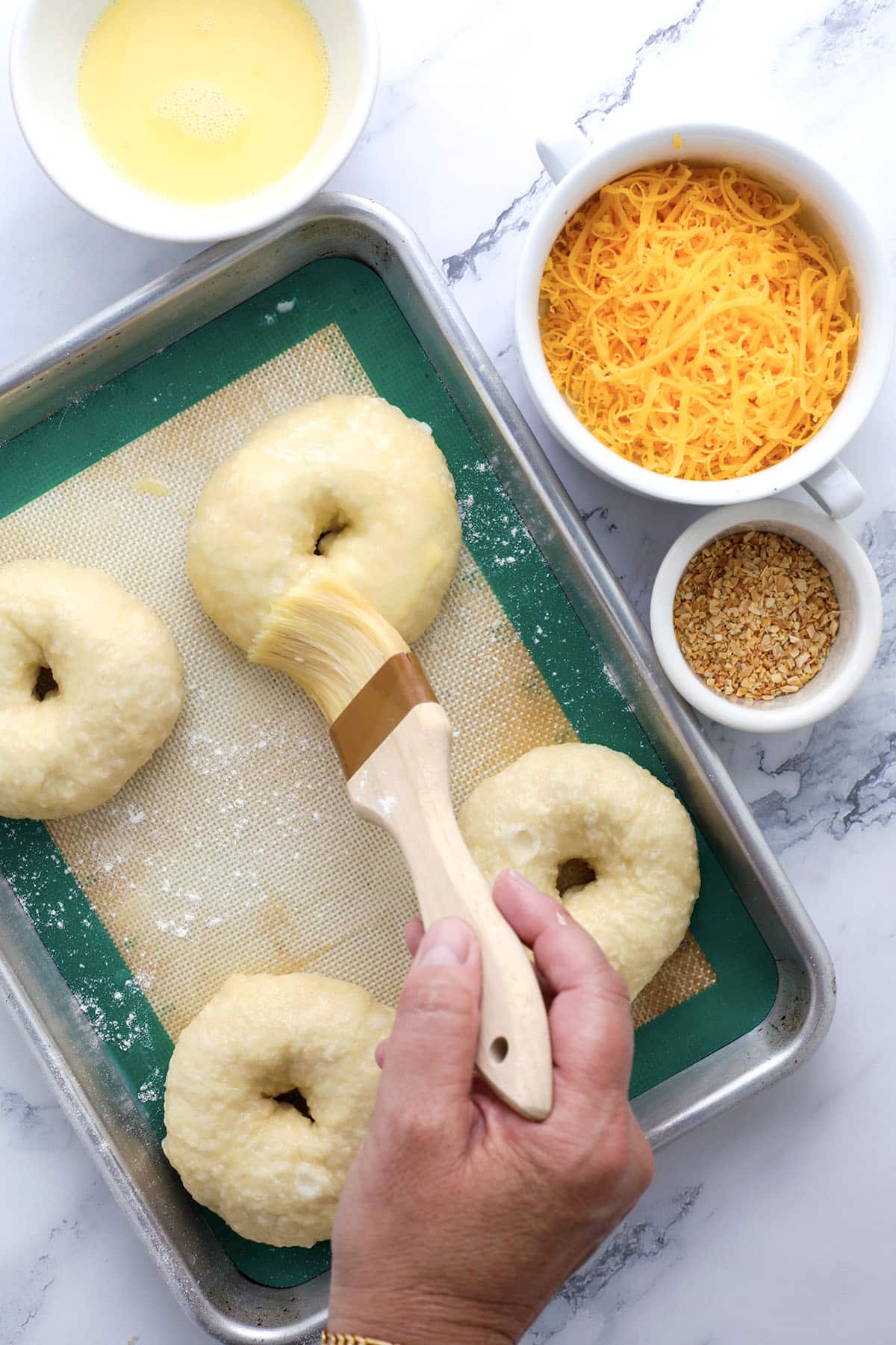 A hand brushes egg wash onto raw bagels on a baking tray—an essential step in how to make bagels at home. Nearby, bowls of shredded cheddar cheese and dried minced onions await for topping these cheesy garlic bagels on a marble countertop.