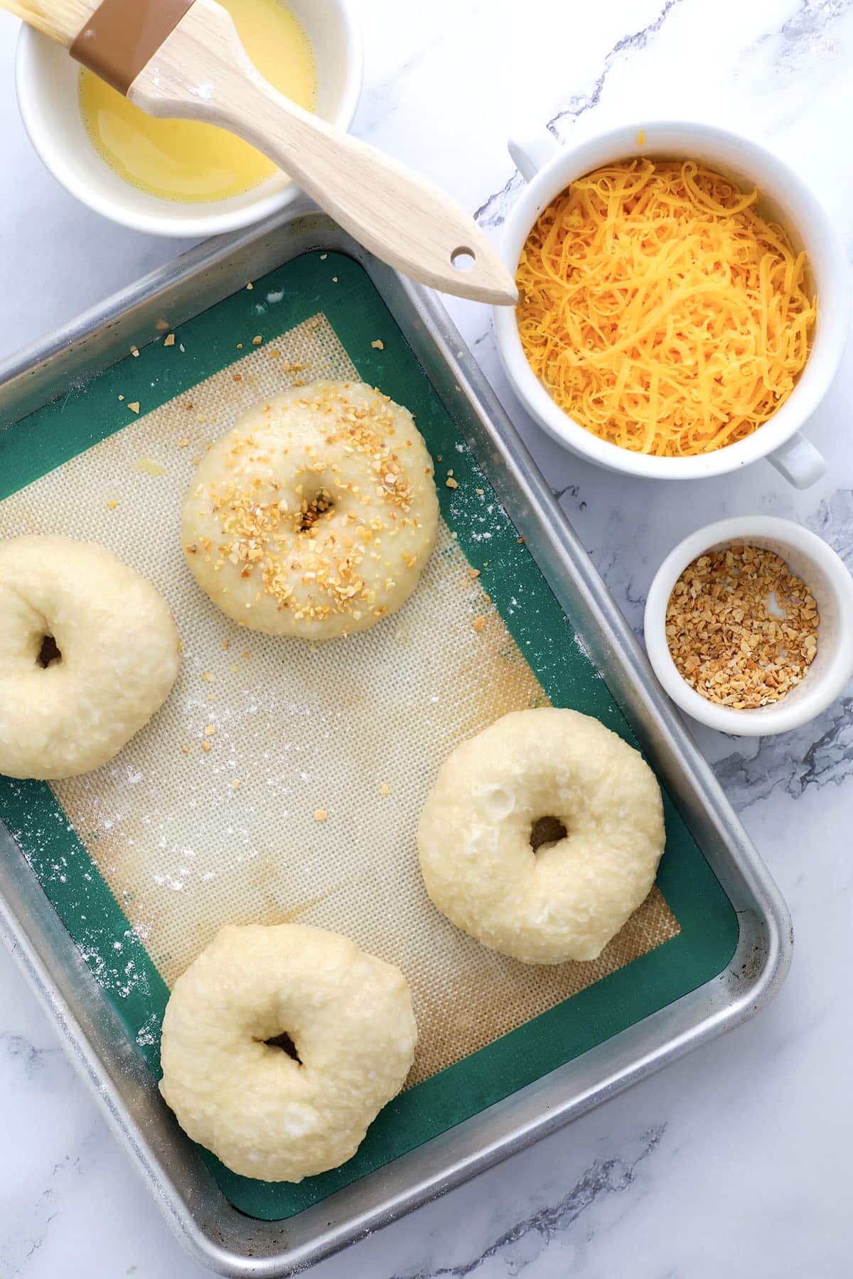 Four unbaked cheddar garlic bagels rest on a baking sheet, one topped with seasoning. Nearby are bowls of shredded cheddar cheese, seasoning, and a brush resting on a bowl of melted butter.