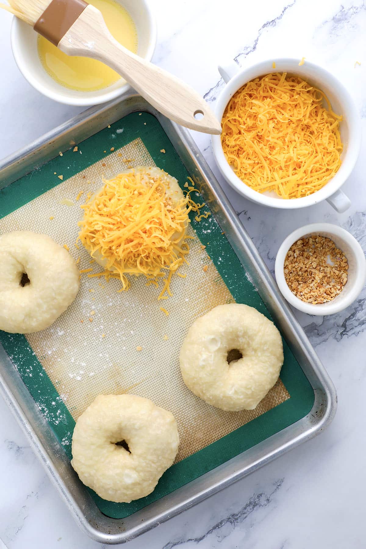 A baking sheet with unbaked cheddar garlic bagels, a bowl of shredded cheddar cheese, a small bowl of seasoning, and a dish of melted butter with a brush, all arranged on a marble countertop—perfect for learning how to make bagels at home.