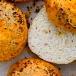 Close-up of golden brown, round cheesy garlic bagels topped with melted cheese and herbs; one is sliced open to reveal a soft, airy interior. Perfect inspiration for how to make bagels at home, shown on a textured baking mat.