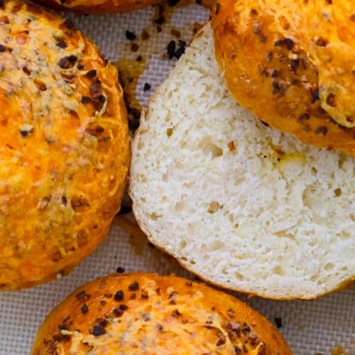 Close-up of golden brown, round cheesy garlic bagels topped with melted cheese and herbs; one is sliced open to reveal a soft, airy interior. Perfect inspiration for how to make bagels at home, shown on a textured baking mat.