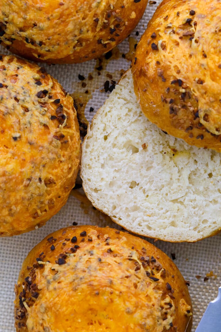 Close-up of golden brown, round cheesy garlic bagels topped with melted cheese and herbs; one is sliced open to reveal a soft, airy interior. Perfect inspiration for how to make bagels at home, shown on a textured baking mat.