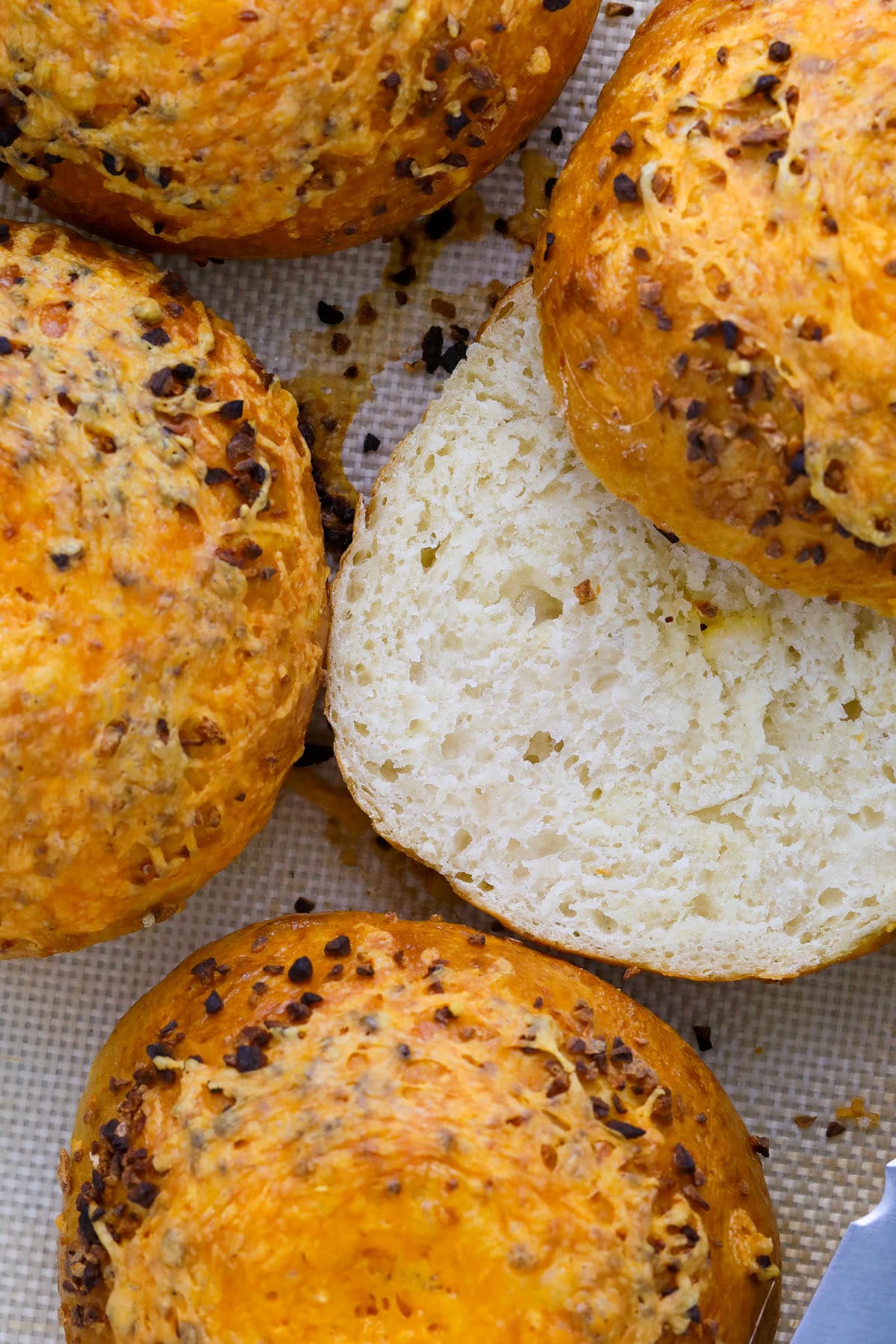 Close-up of golden brown, round cheesy garlic bagels topped with melted cheese and herbs; one is sliced open to reveal a soft, airy interior. Perfect inspiration for how to make bagels at home, shown on a textured baking mat.