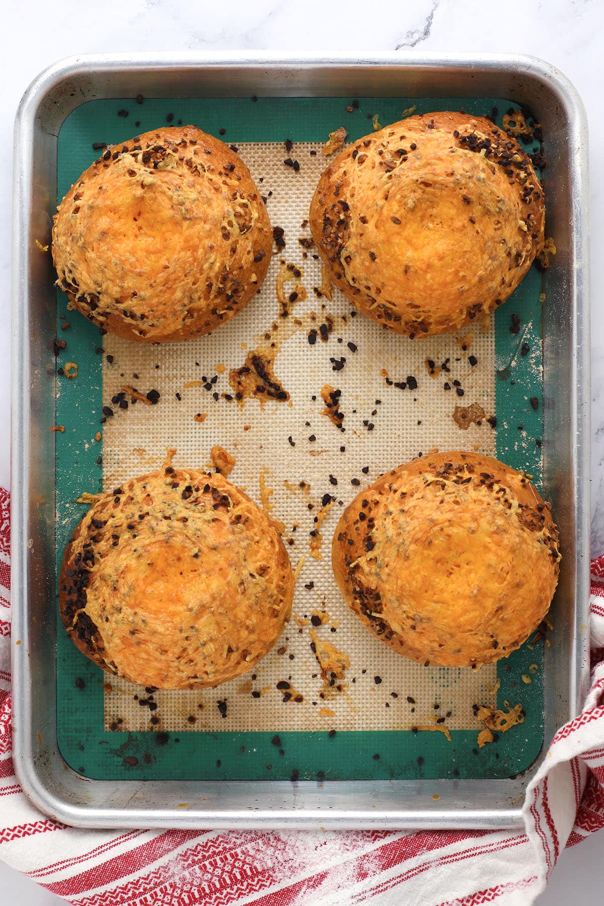 Four golden-brown, round cheesy garlic bagels sit on a silicone baking mat in a metal tray, with scattered bits of melted cheese. A red and white striped towel is placed beside the tray, perfect for anyone learning how to make bagels at home.