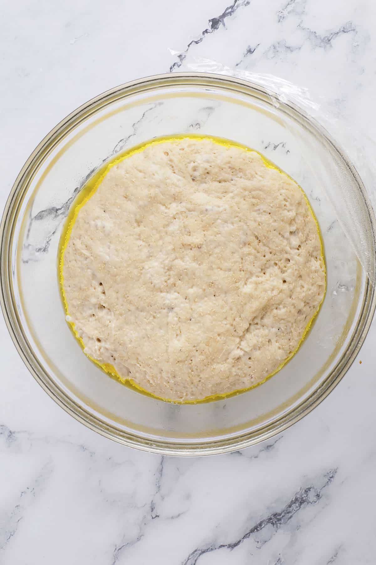 A bowl of risen bread dough, perfect for making cheddar garlic bagels, sits on a marble surface partially covered with plastic wrap and edged with a layer of oil.