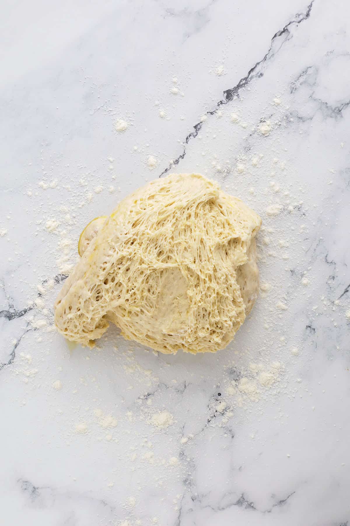 A sticky, airy ball of raw bread dough sprinkled with flour rests on a lightly floured marble surface—perfect for learning how to make bagels or shaping into cheesy garlic bagels.