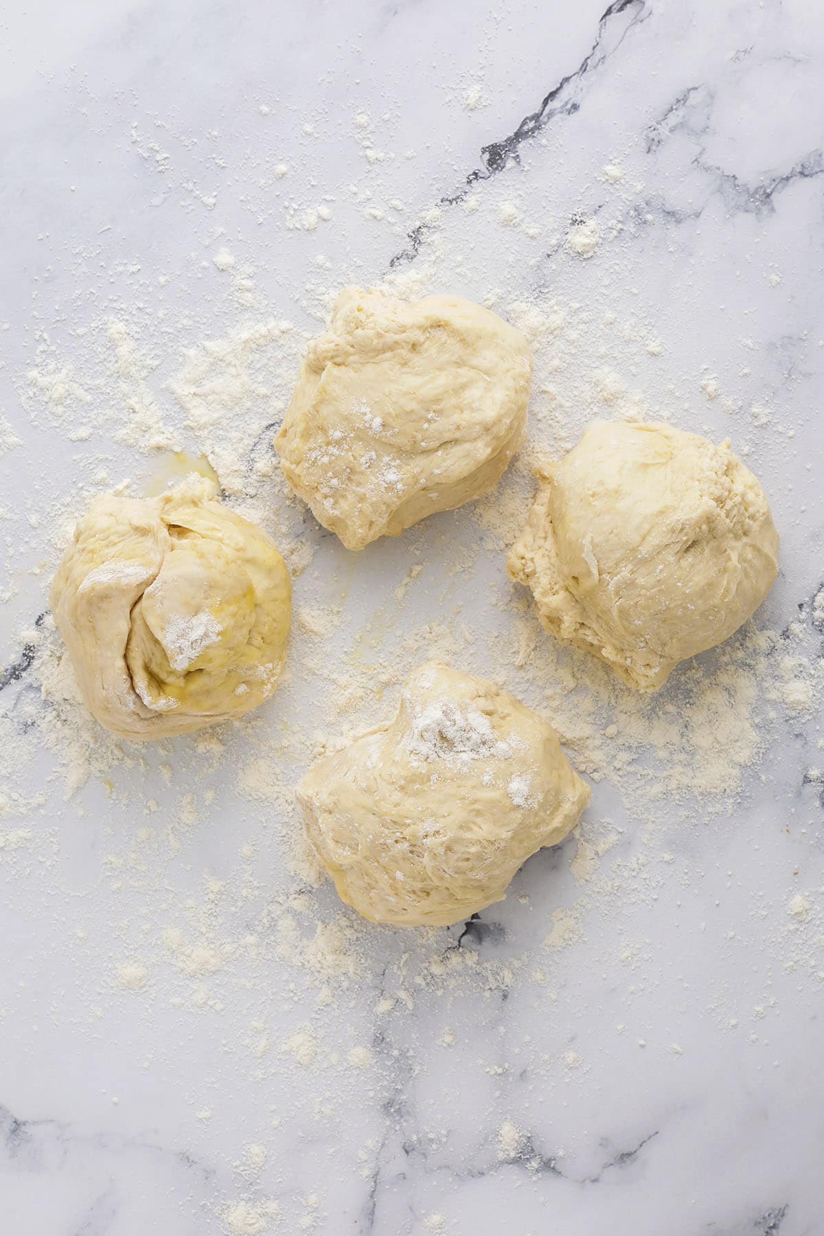 Four portions of raw dough are placed on a floured marble surface, with scattered flour surrounding them—a perfect starting point for learning how to make bagels at home.