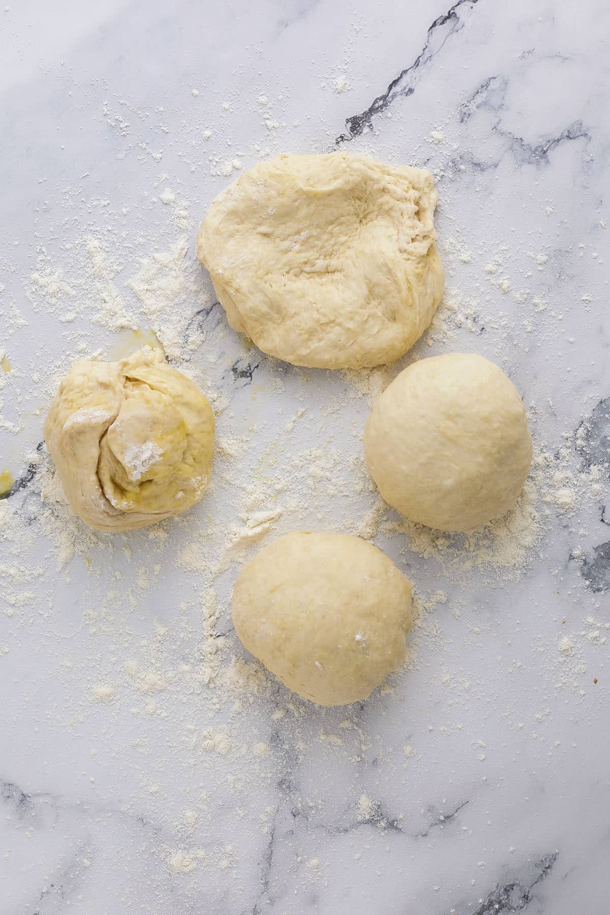 Three round balls and one irregular piece of dough rest on a floured marble surface, perfect for anyone learning how to make bagels at home or shaping cheddar garlic bagels from scratch.