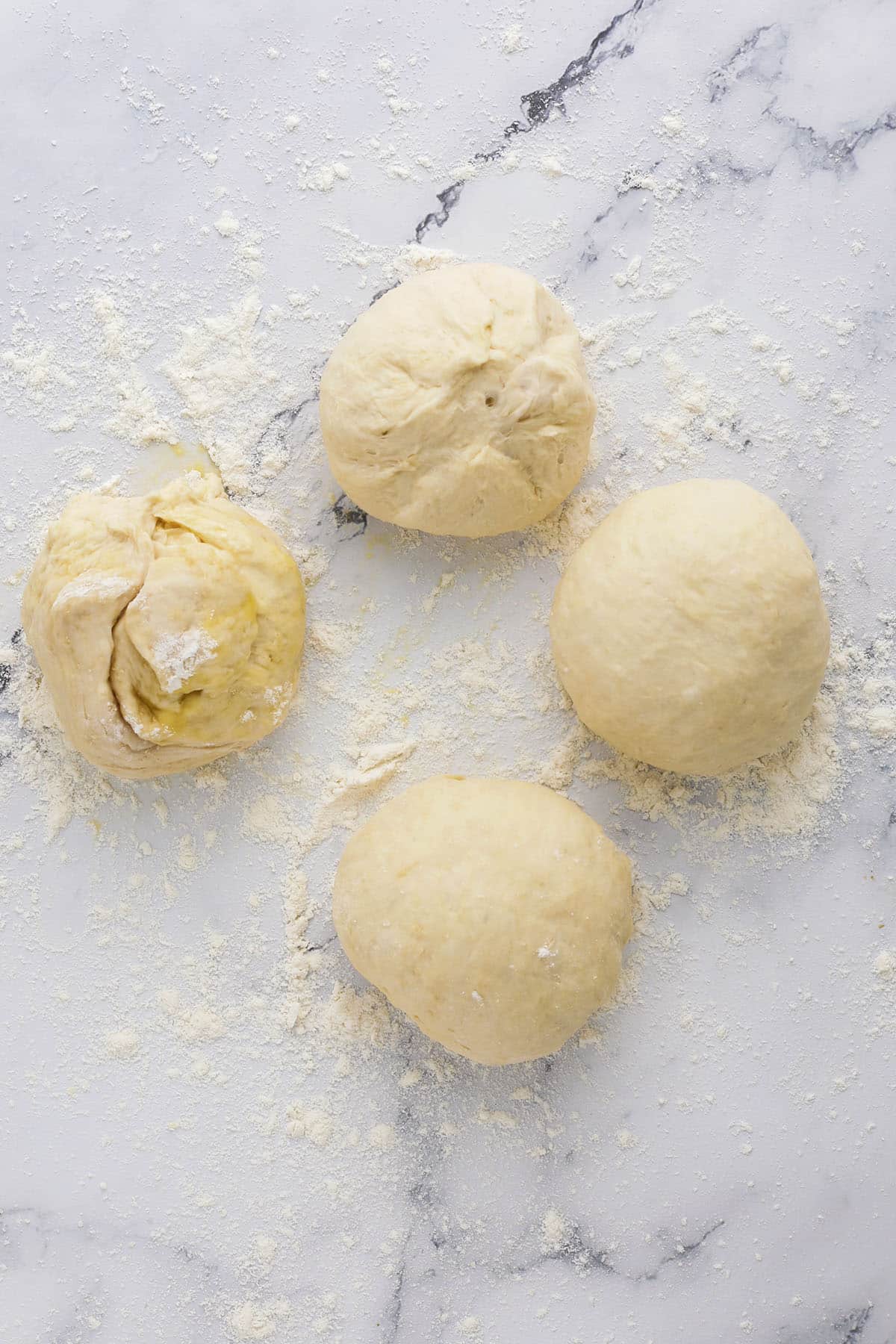 Four balls of pizza dough rest on a floured marble surface, with scattered flour around them. One dough ball is slightly open, showing a soft and airy texture—perfect if you’re curious about how to make bagels at home for a delicious start.