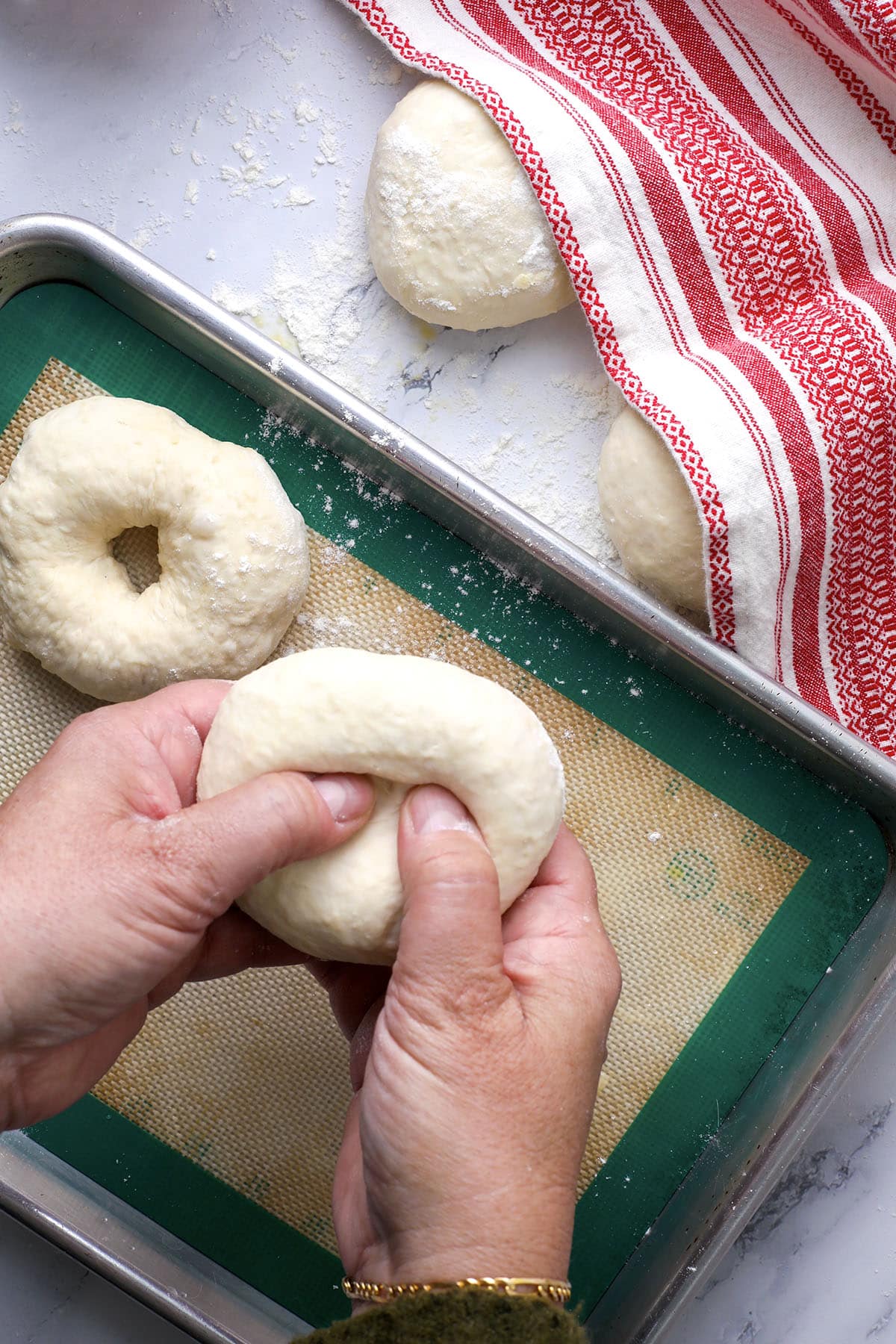 Hands are shaping a round dough ball into a bagel by poking a hole in the center—perfect for those learning how to make bagels at home. A baking tray with a silicone mat, dough balls, flour, and a red-striped towel sit on a light surface.
