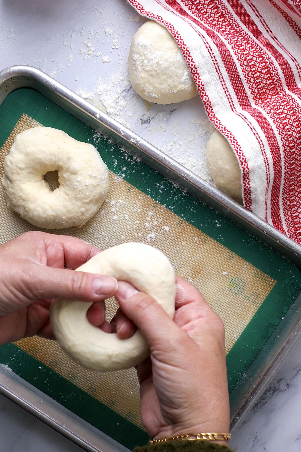 Hands shaping cheesy garlic bagels from dough over a baking sheet with other bagel dough pieces; a red-striped towel and flour are visible on a white countertop—showing how to make bagels at home.