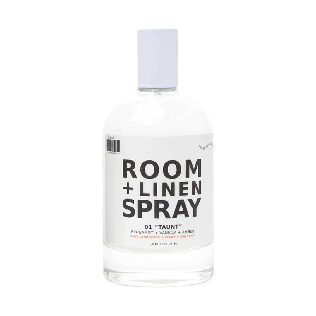 A clear bottle of Room + Linen Spray labeled 01 Taunt with bergamot, vanilla, and amber scents. The bottle has a white cap and minimalist black text design on a white label.