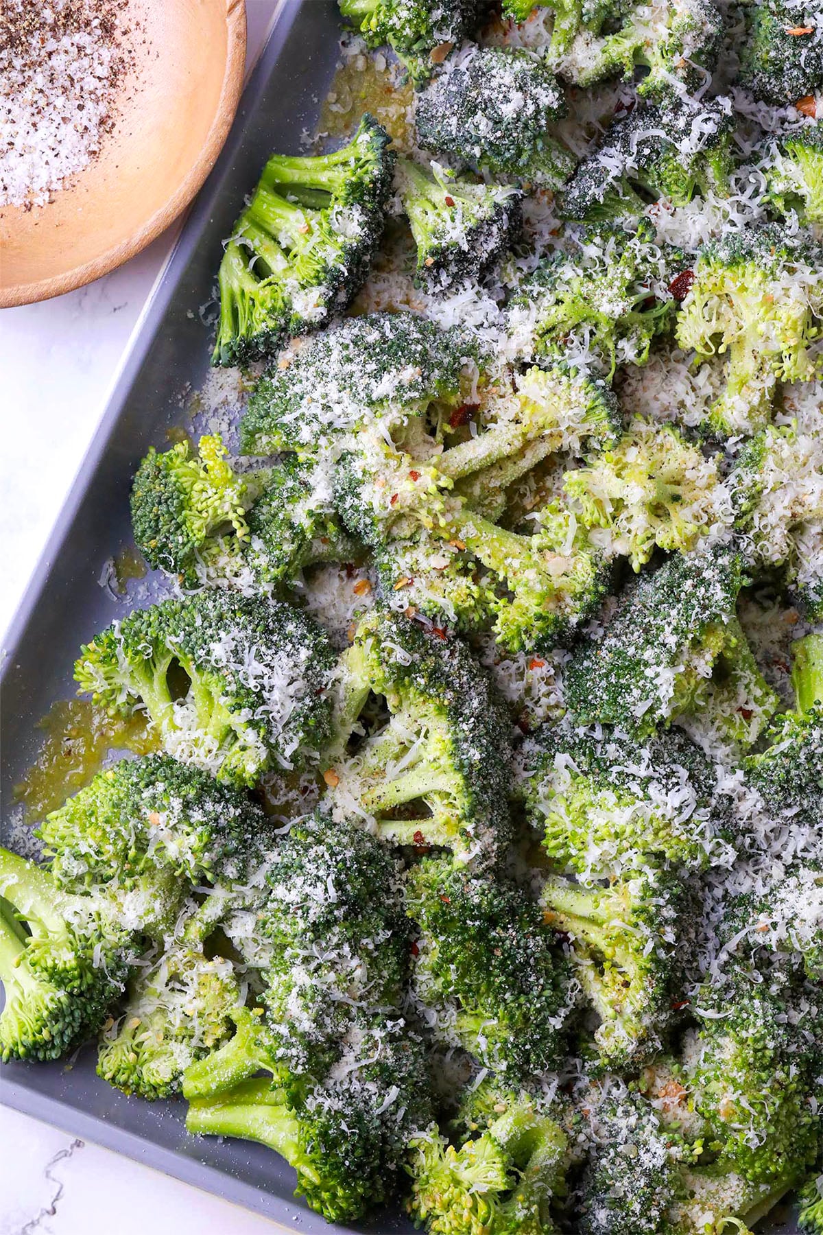 Broccoli florets on a baking tray topped with grated Parmesan cheese and black pepper, ready to be roasted. A wooden bowl with more black pepper is visible in the upper left corner.