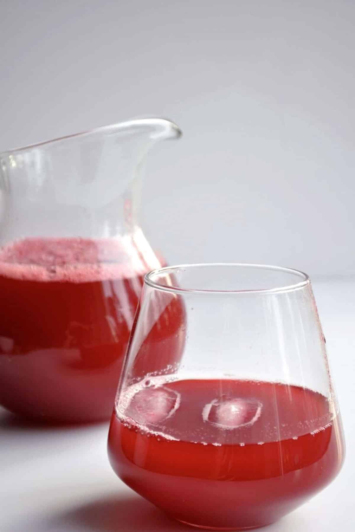 A glass and a pitcher filled with bright red juice sit on a white surface, with light reflecting off the glass and subtle bubbles visible on top of the juice.
