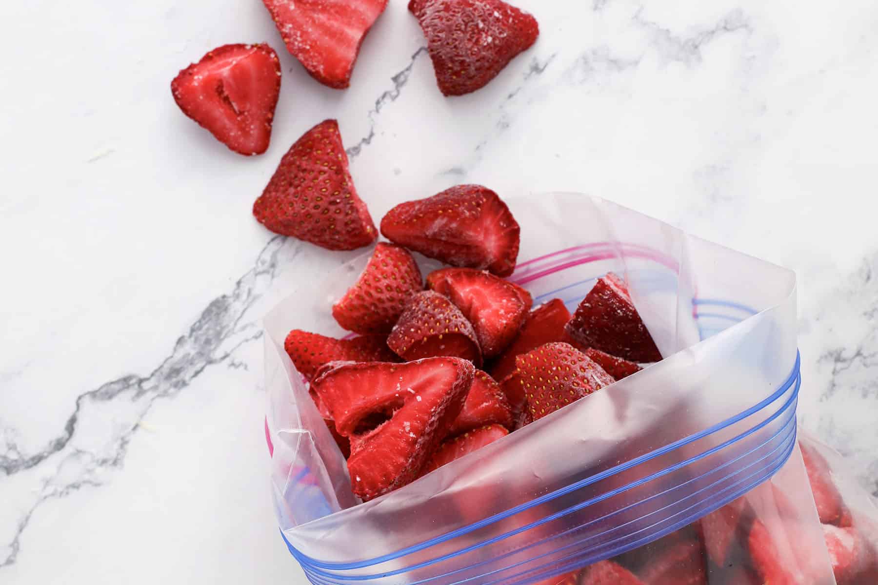Sliced strawberries, some spilling out, are shown in and around a clear resealable plastic bag on a white marble surface.