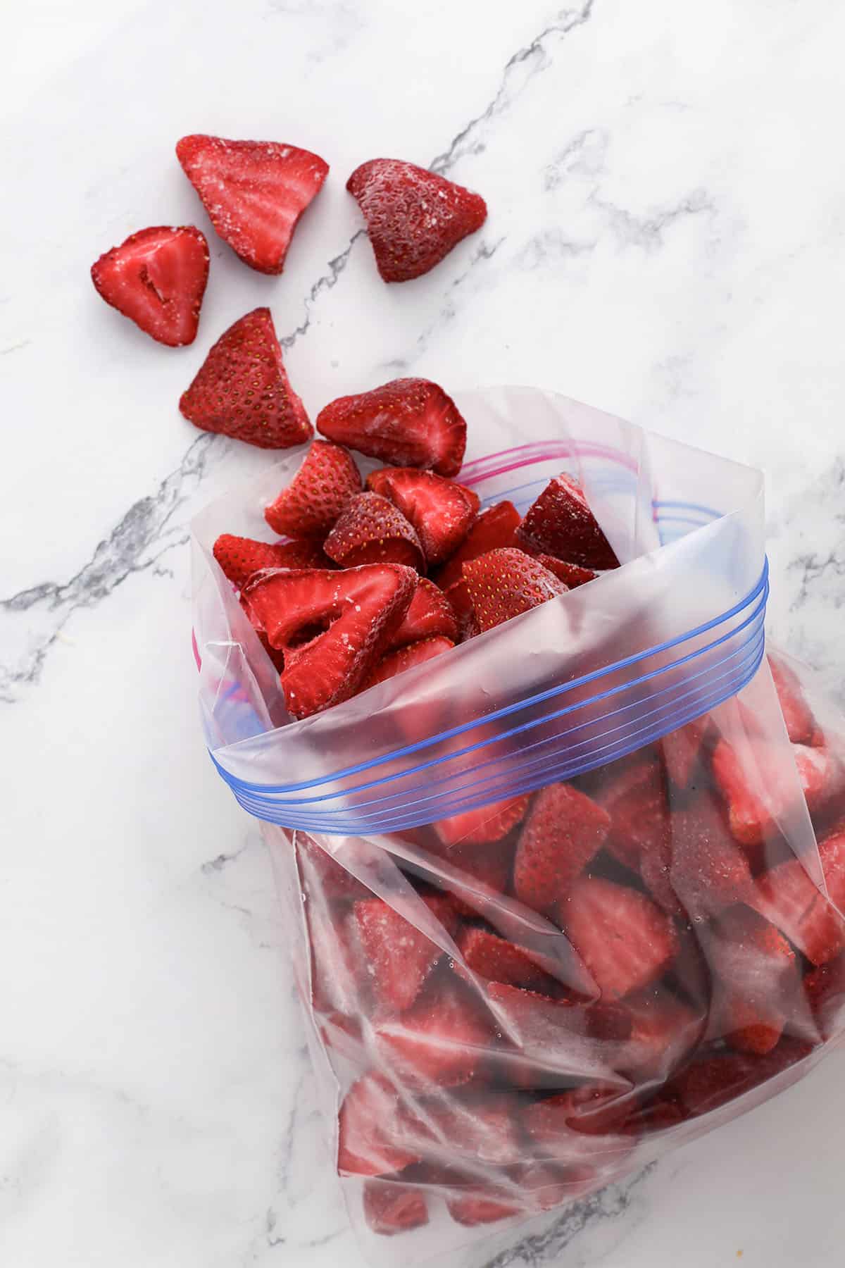 A partially opened plastic ziplock bag filled with frozen strawberry halves sits on a marble surface, with a few strawberries scattered outside the bag.