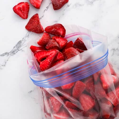 Sliced strawberries in a resealable plastic bag on a marble surface, with a few pieces scattered outside the bag.