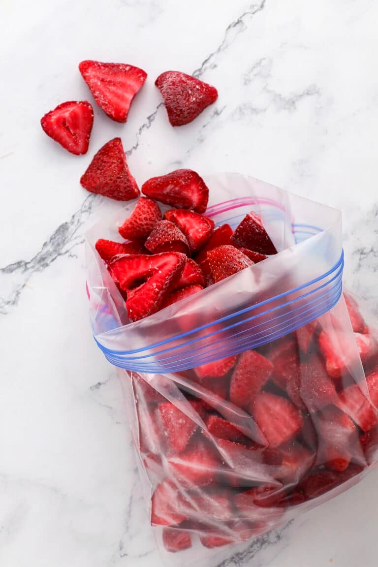 Sliced strawberries in a resealable plastic bag on a marble surface, with a few pieces scattered outside the bag.