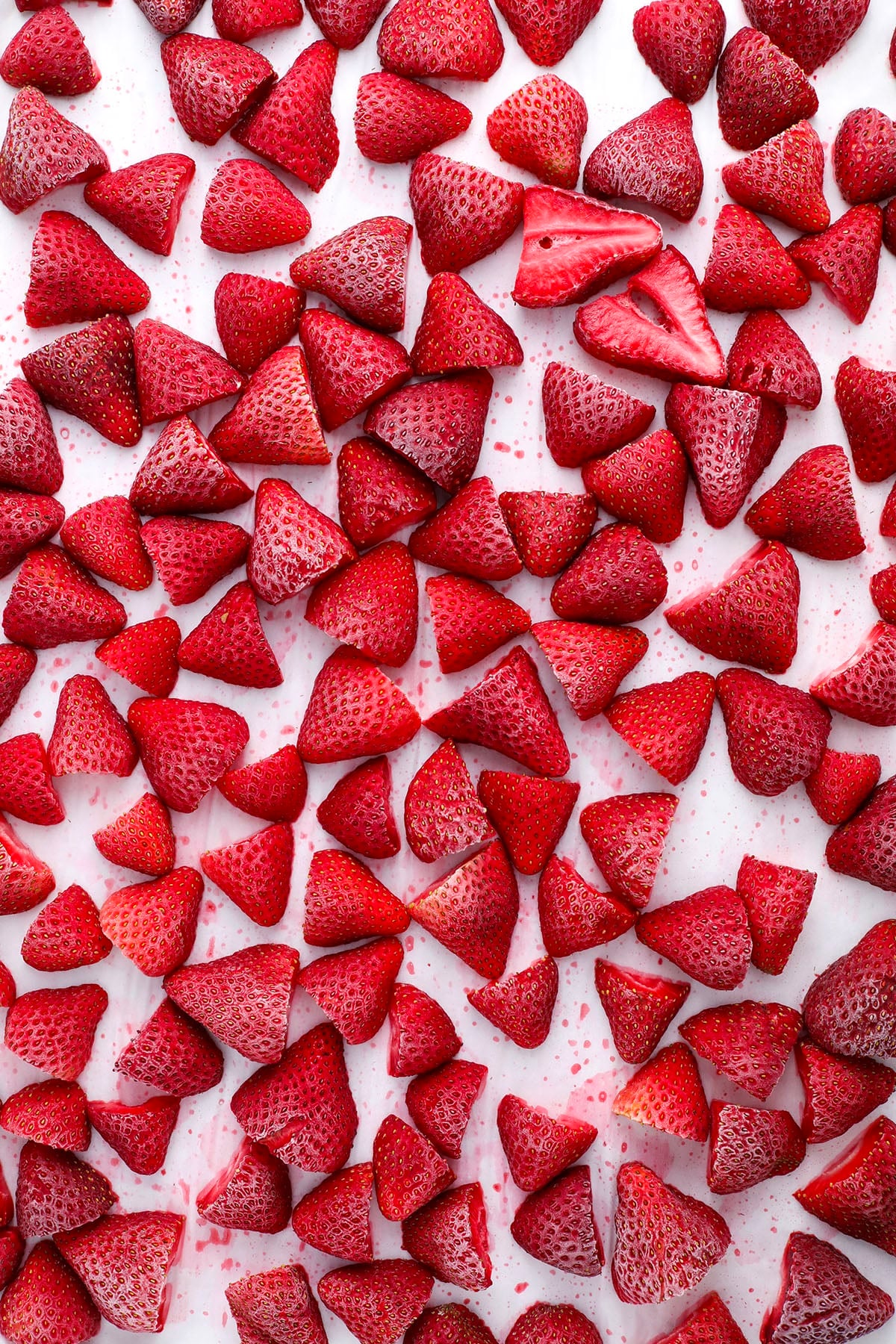 A large number of halved strawberries are scattered evenly across a white surface, with some juice staining the background. The strawberries are bright red and fresh-looking.