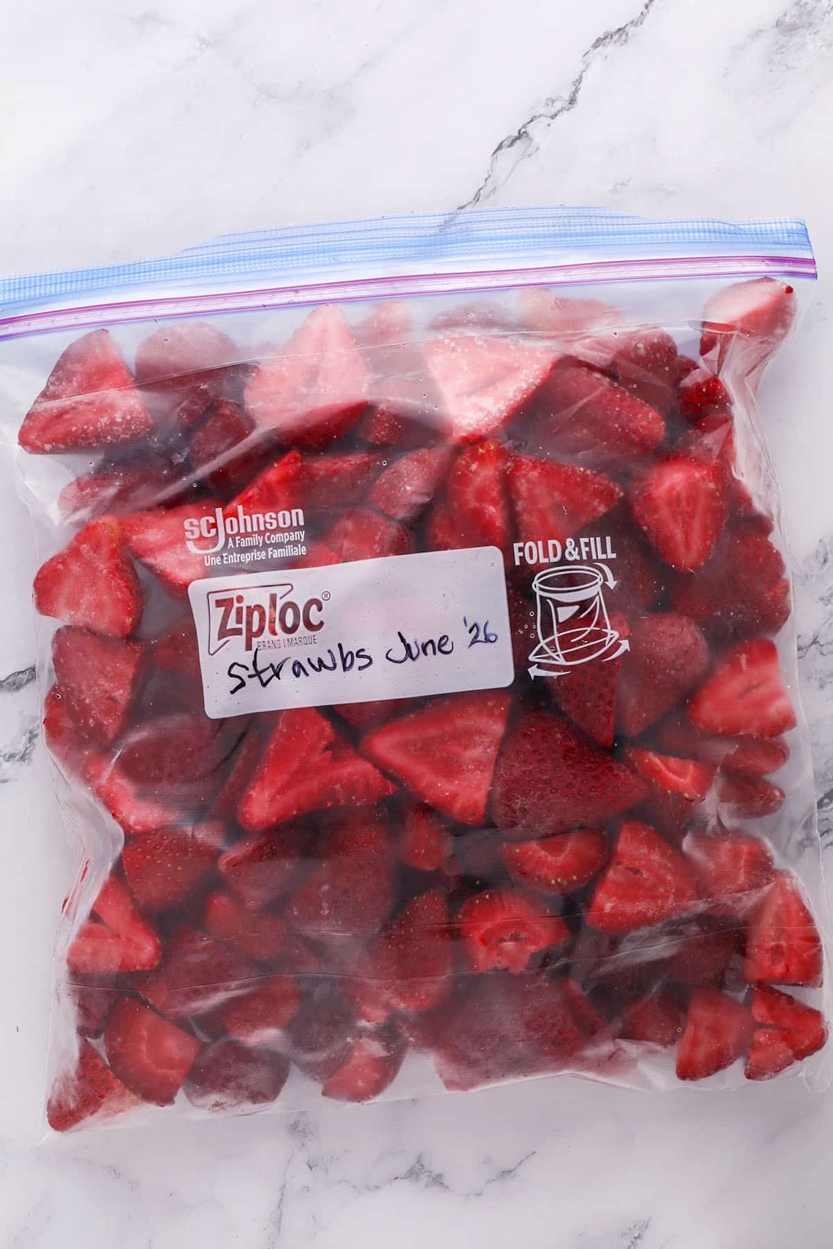 A Ziploc bag filled with sliced strawberries, labeled Strawbs June 26, sits on a white marble surface.
