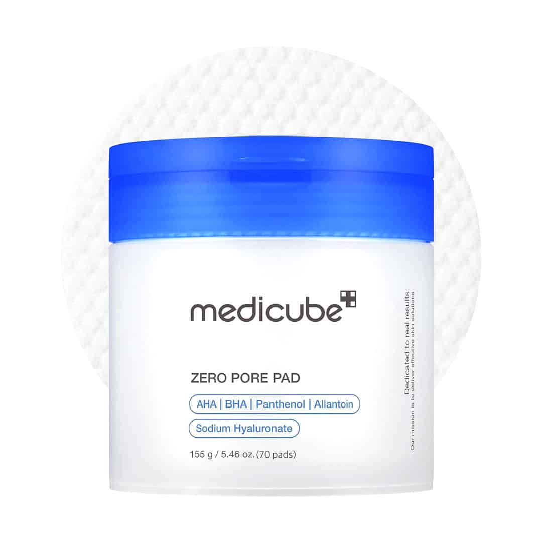 A white jar with a blue lid labeled medicube ZERO PORE PAD stands in front of a round cotton pad. Key ingredients and product details are listed on the front of the jar.