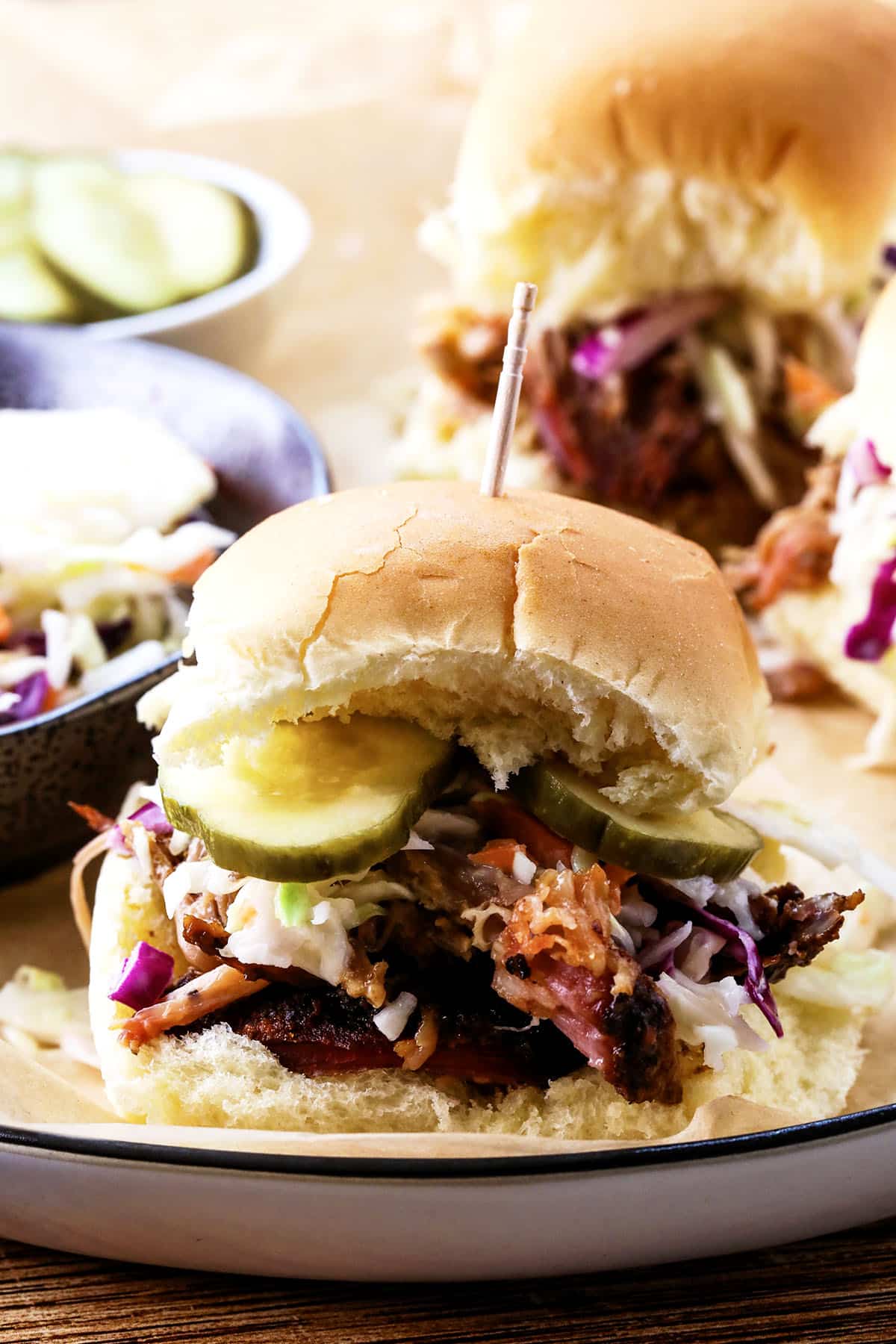 A pulled pork slider topped with coleslaw and pickle slices on a soft bun, secured with a toothpick, is served on a plate with more sliders and a bowl of pickles in the background.