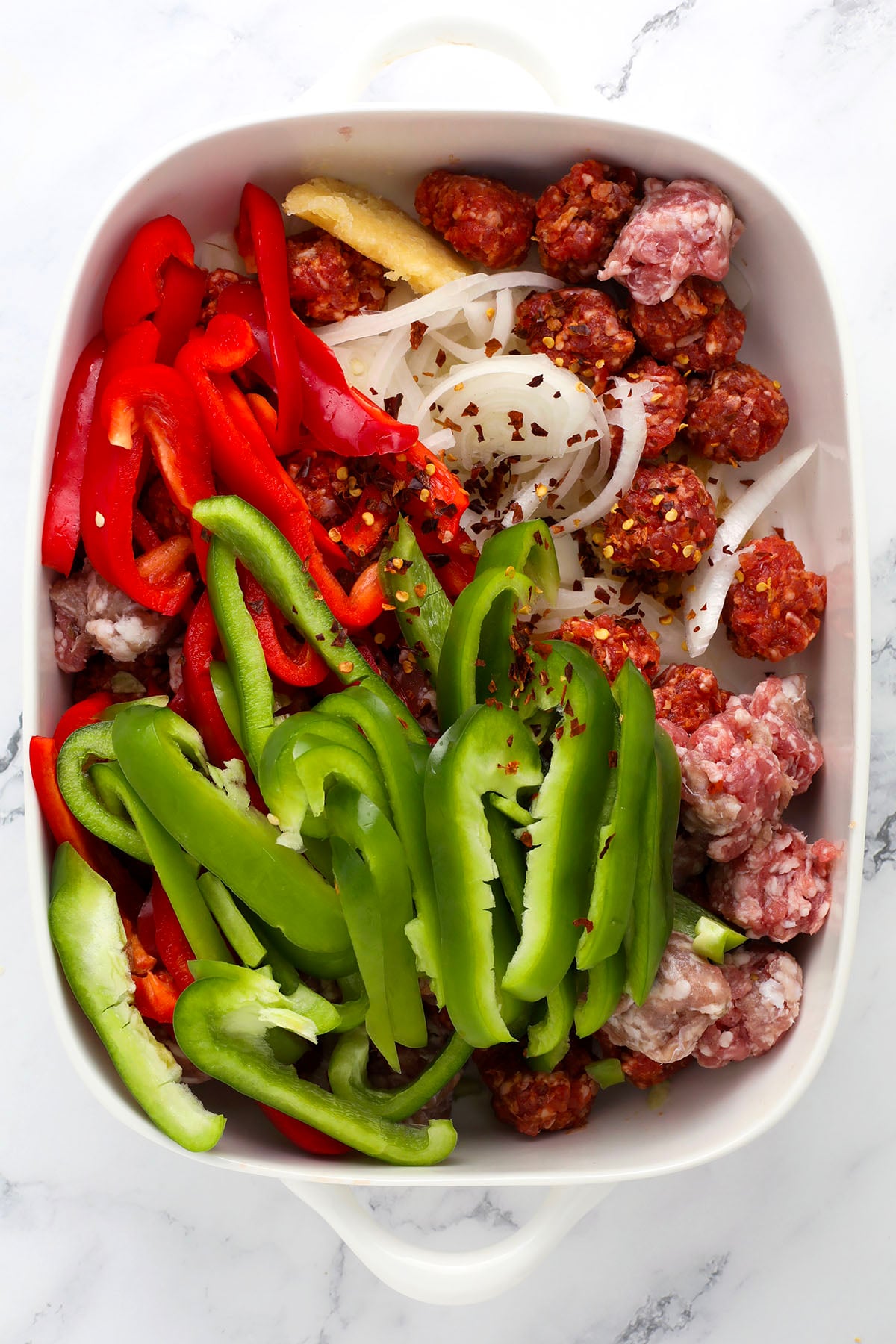 A white baking dish filled with sliced green and red bell peppers, sliced onions, and raw sausage pieces, all topped with red pepper flakes, on a marble surface.